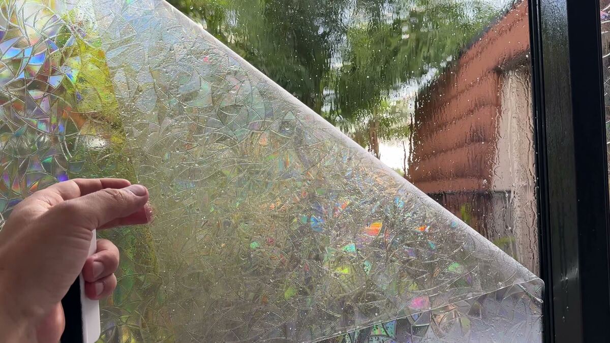 Hands peeling the liner off a sheet of rainbow window film next to a window.